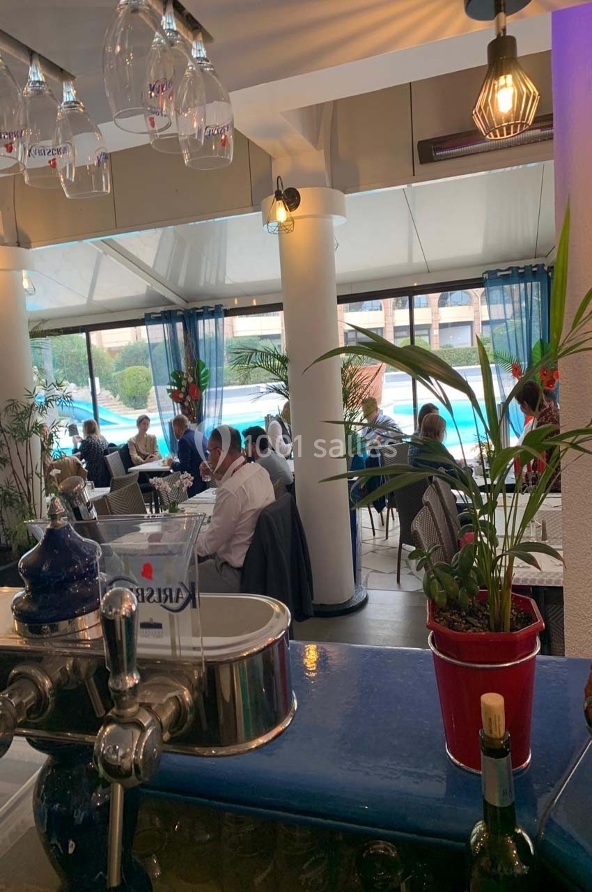 Salle de restaurant lumineuse avec des clients attablés près de grandes baies vitrées donnant sur une piscine.