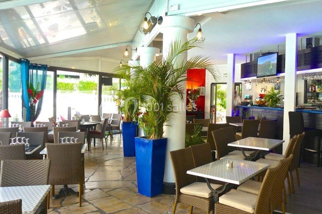 Salle de restaurant lumineuse avec mobilier en rotin, plantes décoratives et bar en arrière-plan.