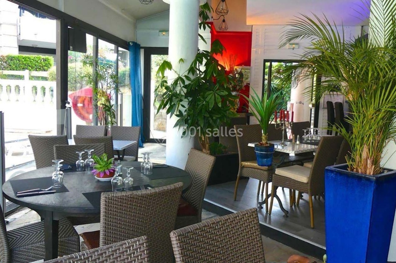 Salle de restaurant lumineuse avec tables en osier, plantes décoratives et grandes baies vitrées donnant sur l'extérieur.