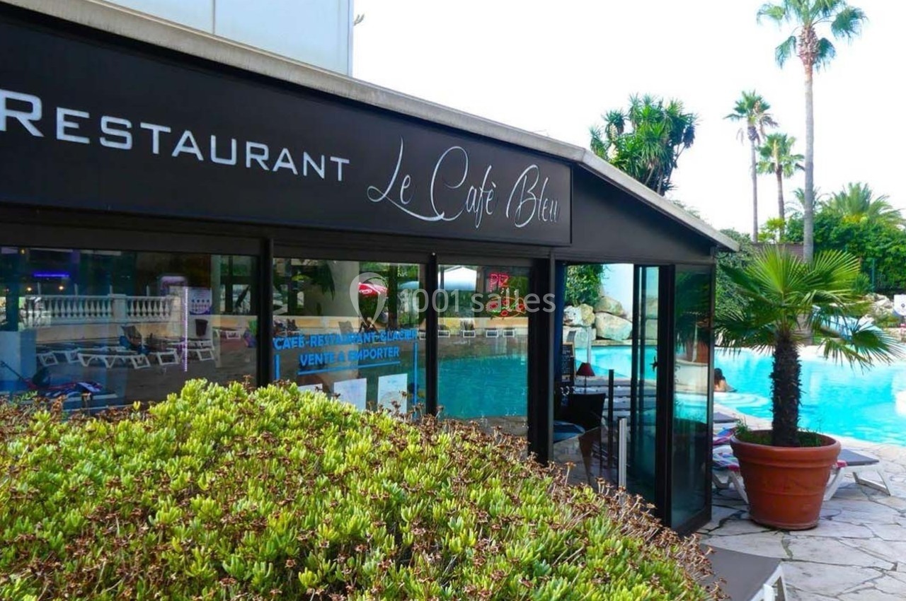 Façade vitrée d'un restaurant avec terrasse donnant sur une piscine entourée de végétation et de palmiers.