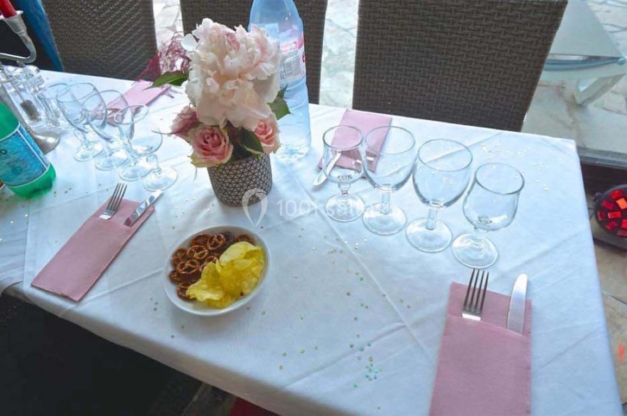 Table dressée avec nappes blanches, serviettes roses, verres alignés, fleurs au centre et petites assiettes d'apéritifs.