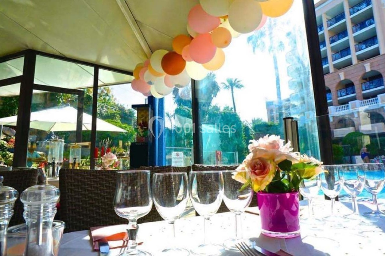 Table dressée avec des verres à pied et un bouquet de fleurs, sous une décoration de ballons près d'une piscine en extérieur.