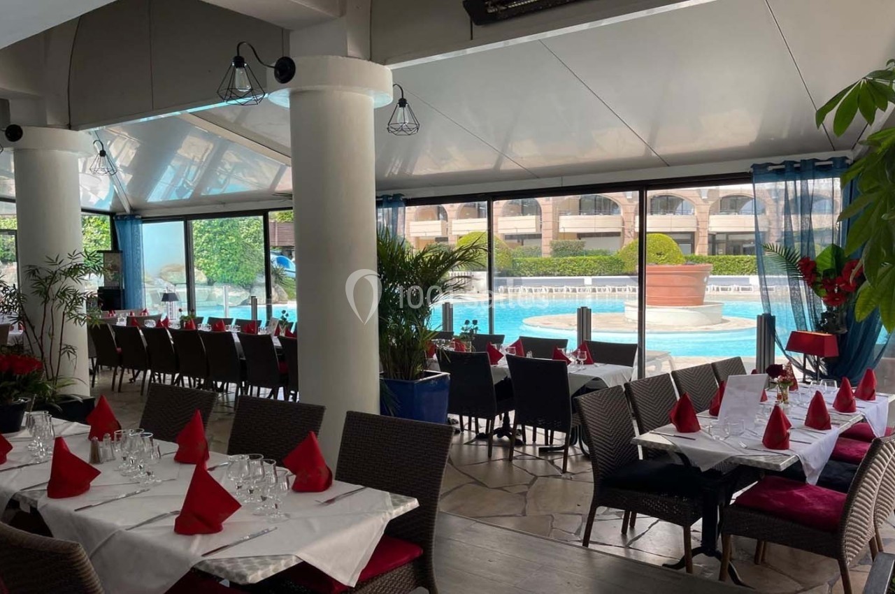 Salle de restaurant lumineuse avec tables dressées, nappes blanches et serviettes rouges, vue sur une piscine extérieure.