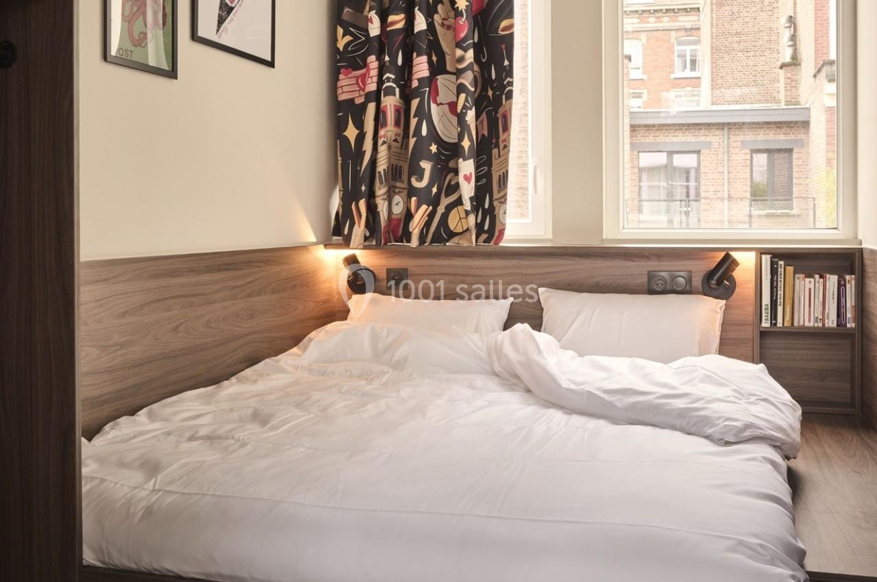 Lit double avec draps blancs dans une chambre lumineuse, décorée de rideaux colorés et d'étagères avec des livres.