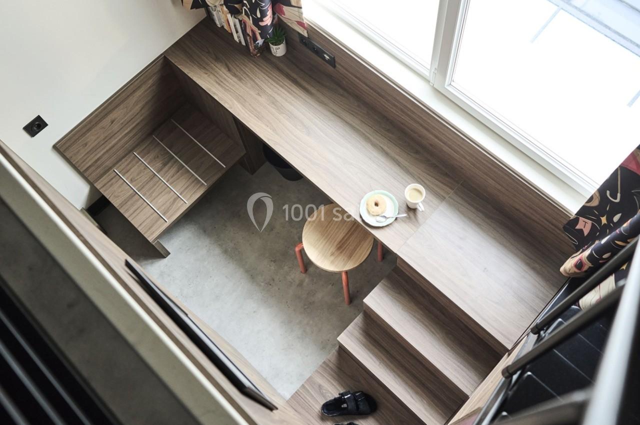 Vue d'en haut d'un espace de travail compact avec bureau en bois, tabouret, tasse de café et assiette sur une mezzanine.