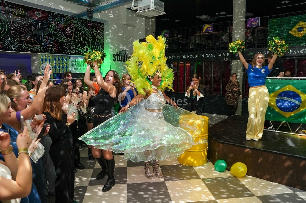Danseuse en tenue brésilienne entourée de spectateurs et de pom-pom girls dans une ambiance festive en intérieur.