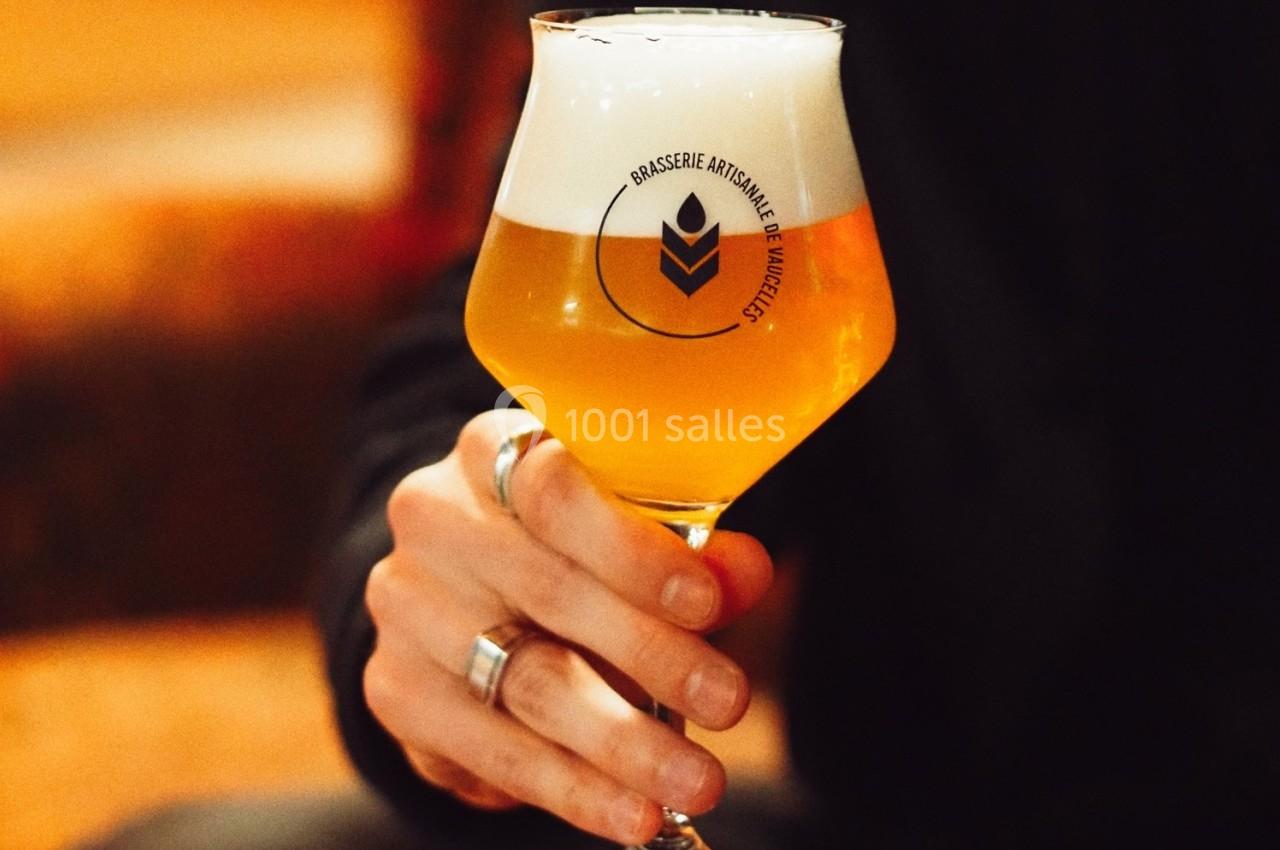 Main tenant un verre de bière artisanale dorée avec une mousse blanche, logo de brasserie visible sur le verre.