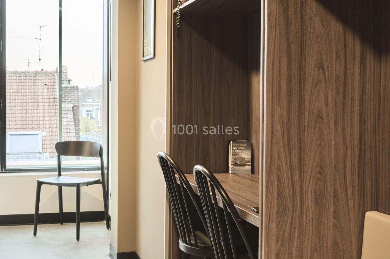 Bureau en bois avec deux chaises noires, près d'une fenêtre offrant une vue sur des toits et un ciel dégagé.
