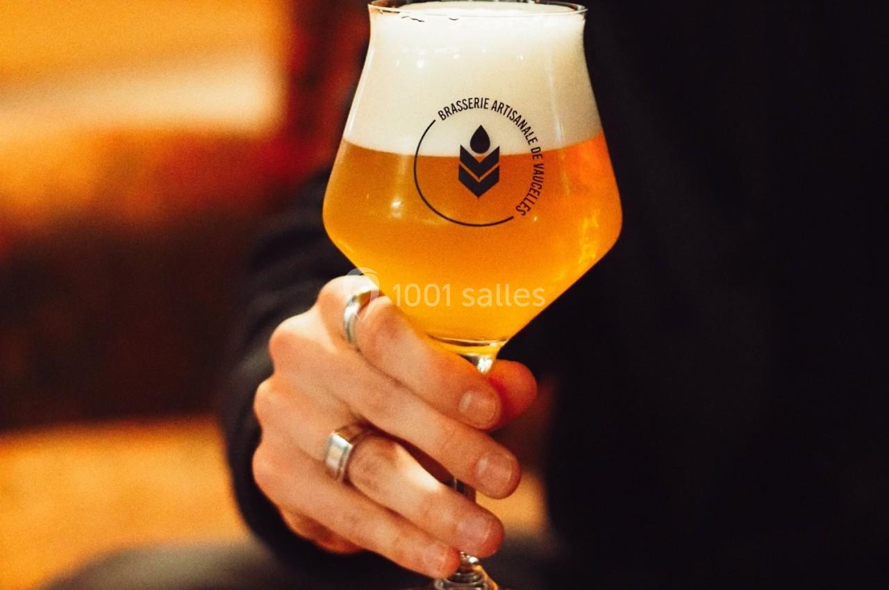 Main tenant un verre de bière artisanale dorée avec une mousse blanche, logo visible sur le verre.