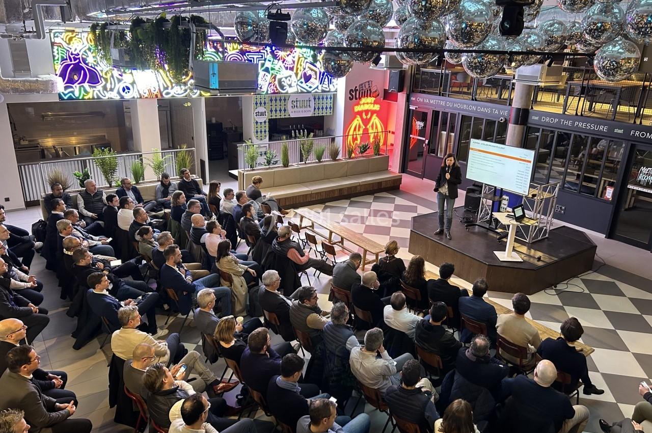 Conférence en intérieur avec un orateur sur scène face à un public assis, dans un espace moderne et lumineux.