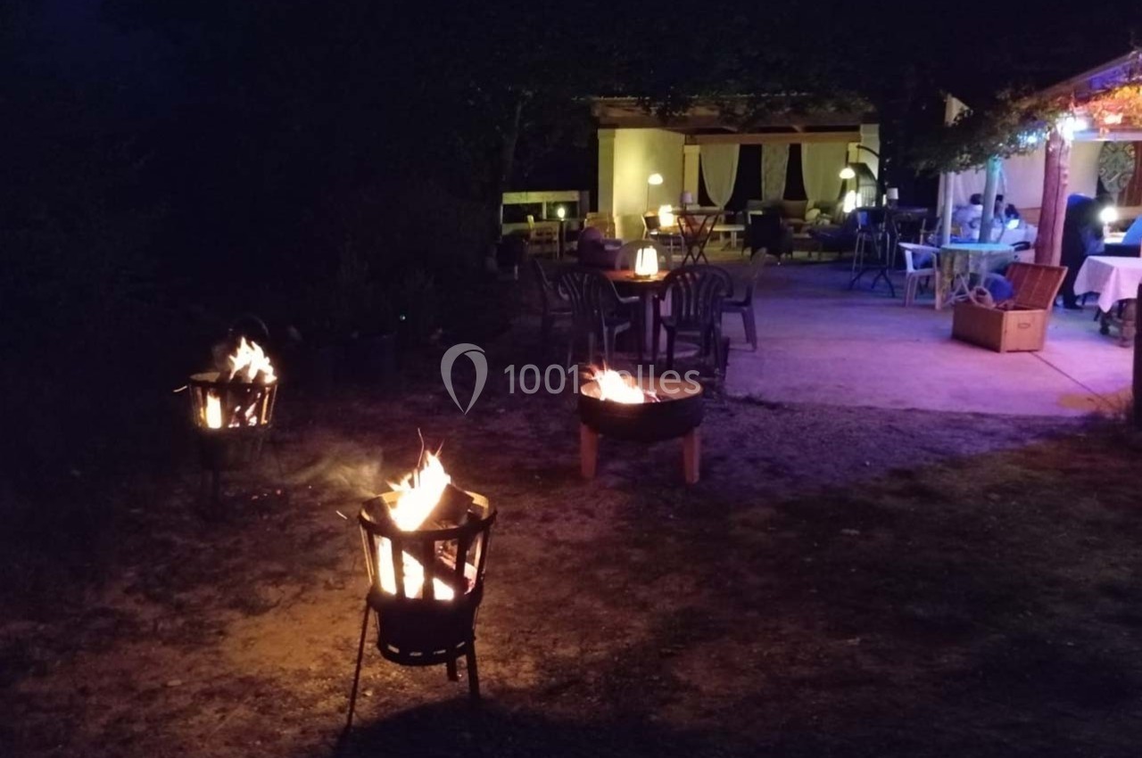 Terrasse éclairée de nuit avec des braseros allumés et des tables disposées sous une pergola.