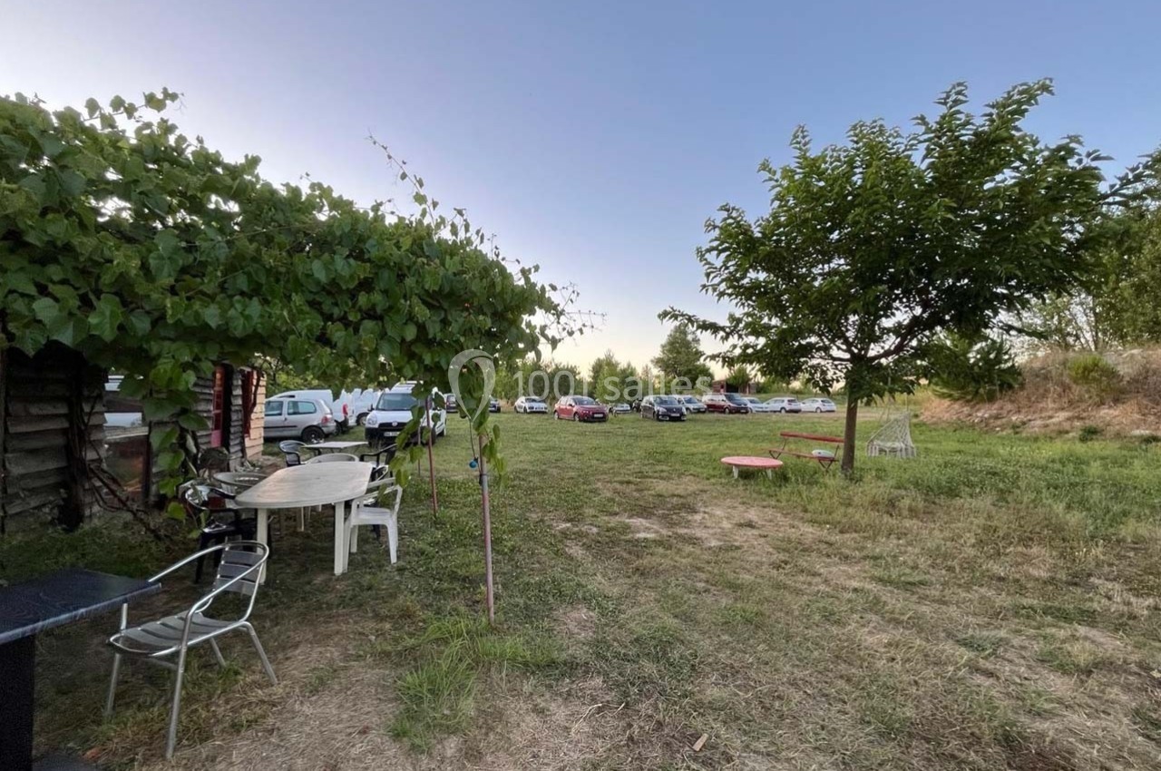 Espace extérieur avec des tables, des chaises, des arbres et des voitures garées en arrière-plan sur un terrain dégagé.