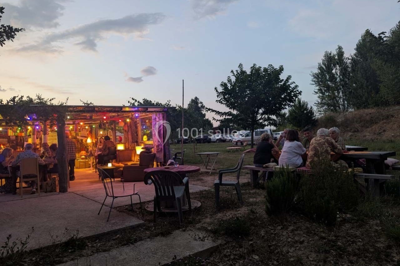 Terrasse extérieure en soirée avec des tables occupées, éclairée par des lumières colorées, entourée de végétation.