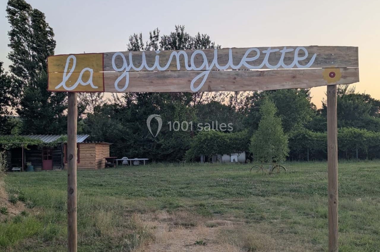 Panneau en bois marqué ’la guinguette’ dans un cadre champêtre avec arbres et abri en arrière-plan.