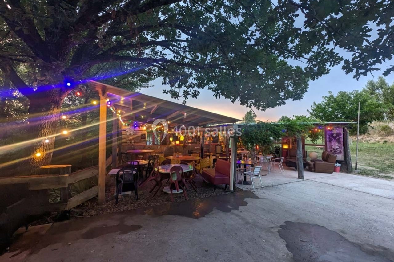 Terrasse extérieure éclairée par des guirlandes lumineuses colorées, entourée de verdure et aménagée avec des tables et…