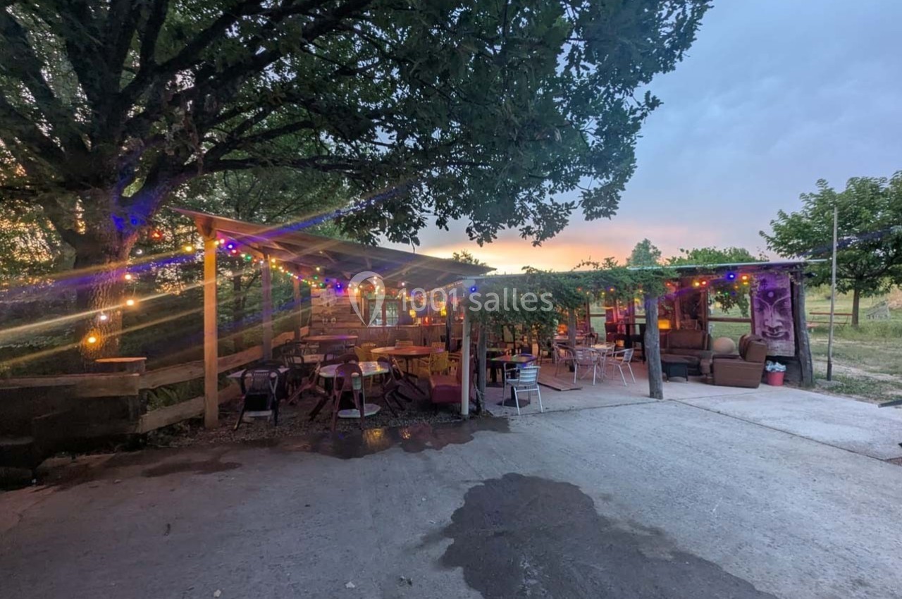 Terrasse extérieure décorée de guirlandes lumineuses colorées, entourée d'arbres et de végétation au coucher du soleil.