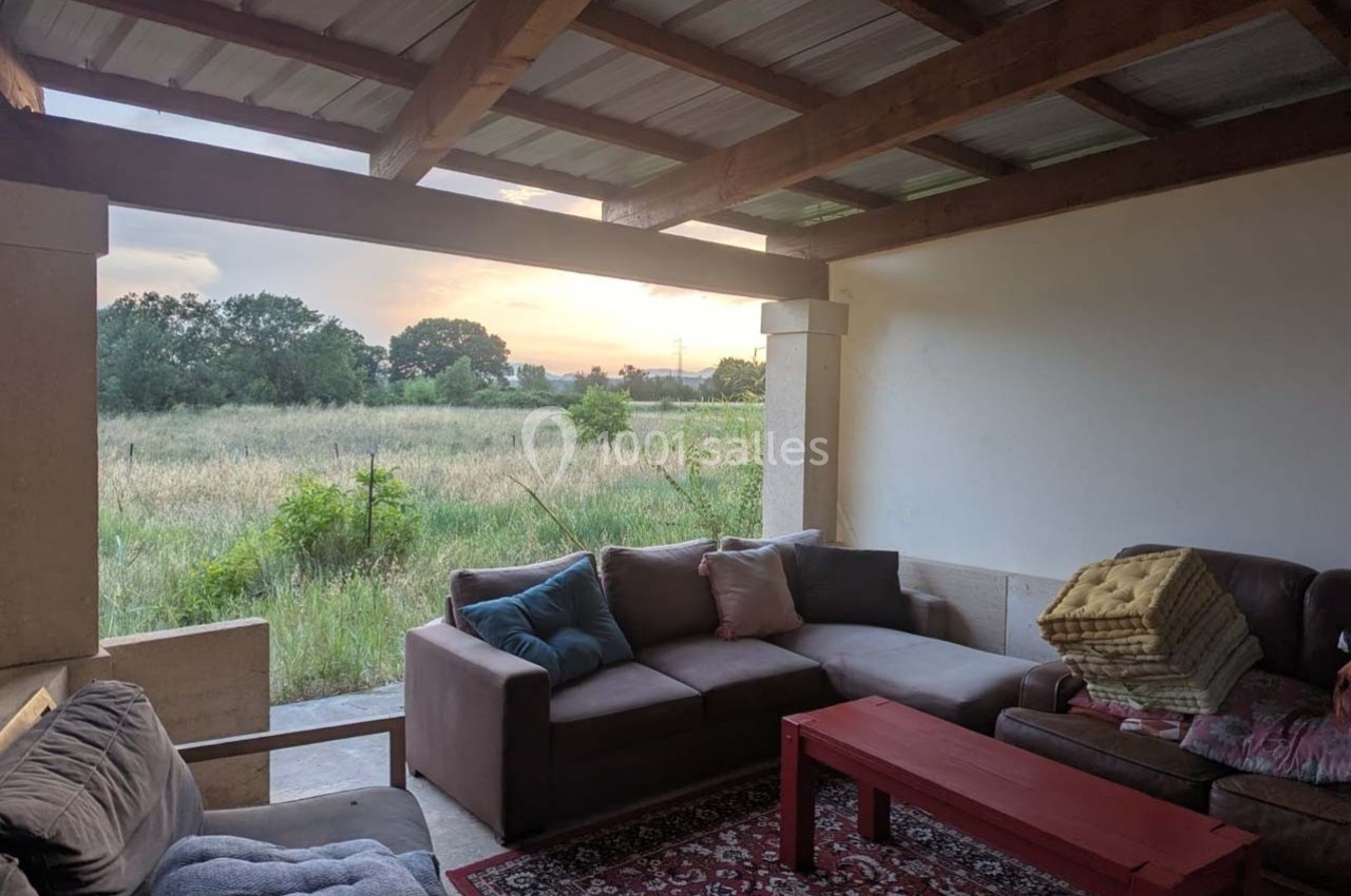 Terrasse couverte avec canapé, table basse rouge, tapis et vue sur un champ au coucher du soleil.