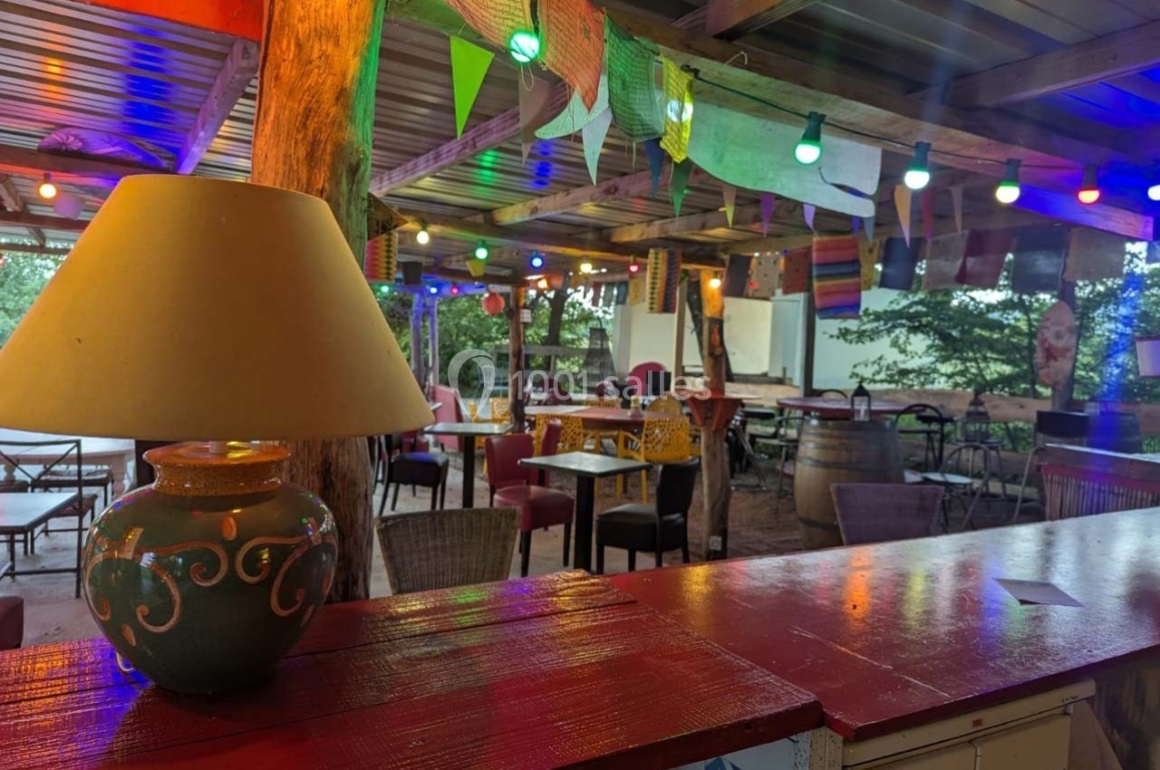 Terrasse couverte décorée de guirlandes lumineuses colorées, avec tables, chaises et une lampe posée sur un comptoir rouge.