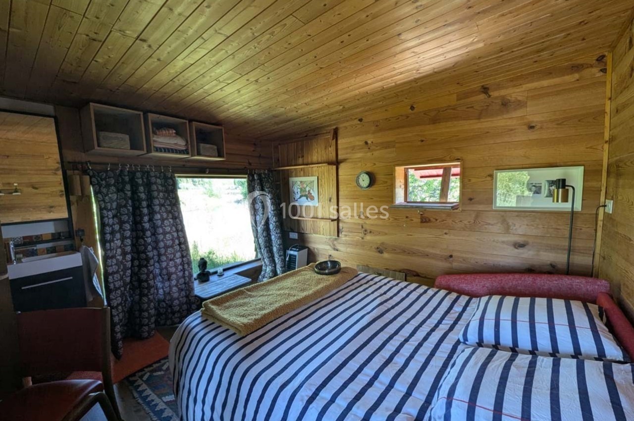 Chambre en bois avec lit à rayures, fenêtres donnant sur l'extérieur, étagères et rideaux sombres.