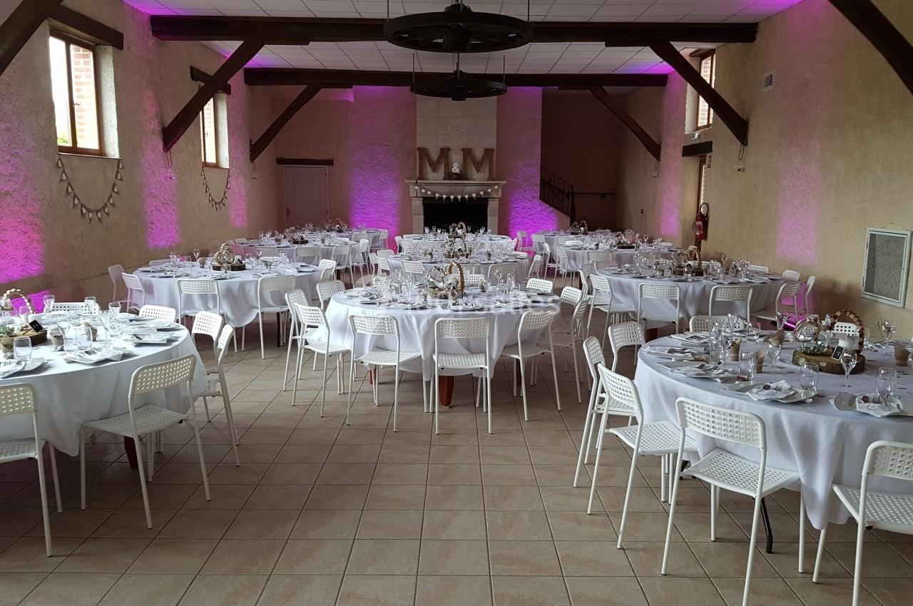 Salle de réception décorée avec des tables rondes dressées, nappes blanches et éclairage d’ambiance rose.