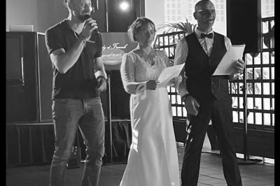 Trois personnes debout, dont une en robe de mariée, parlent ou lisent dans une salle éclairée.