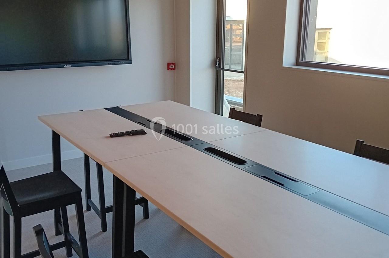 Salle de réunion avec table rectangulaire, chaises noires, écran mural et fenêtre laissant entrer la lumière naturelle.