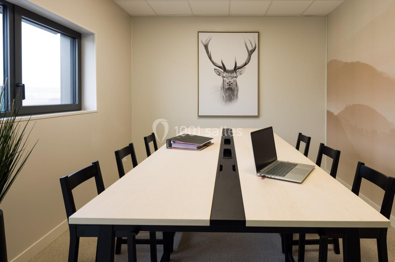 Salle de réunion avec une table rectangulaire, des chaises noires, un ordinateur portable et un tableau représentant un cerf.