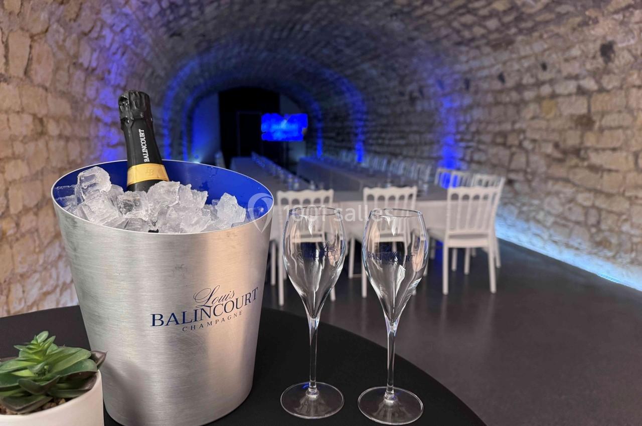 Seau à champagne avec bouteille sur glace, deux flûtes vides et une plante, dans une salle voûtée éclairée en bleu.