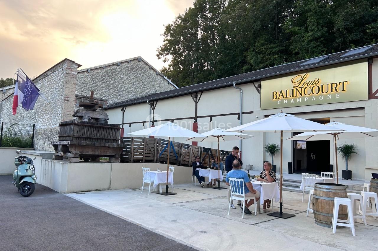 Terrasse extérieure d'une maison de champagne avec tables, parasols, pressoir ancien et façade éclairée au crépuscule.