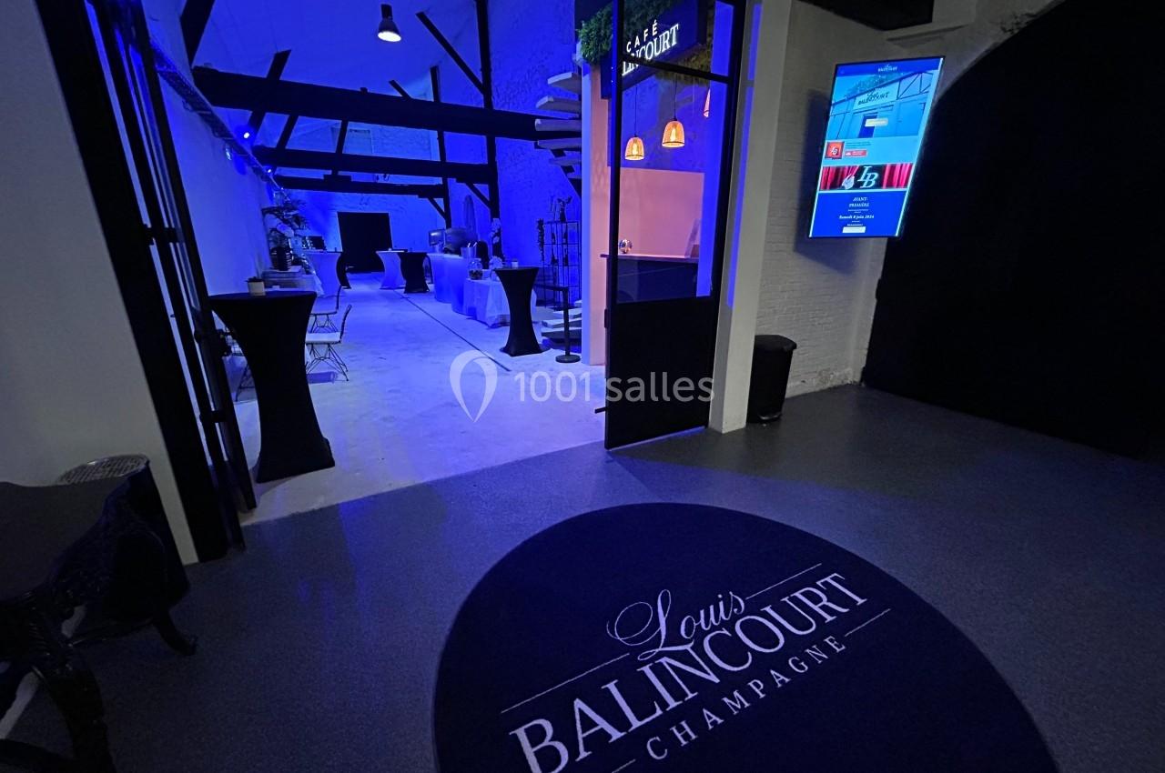 Entrée d'une salle de réception moderne éclairée en bleu, avec tables hautes et tapis marqué ’Louis Balincourt Champagne’.