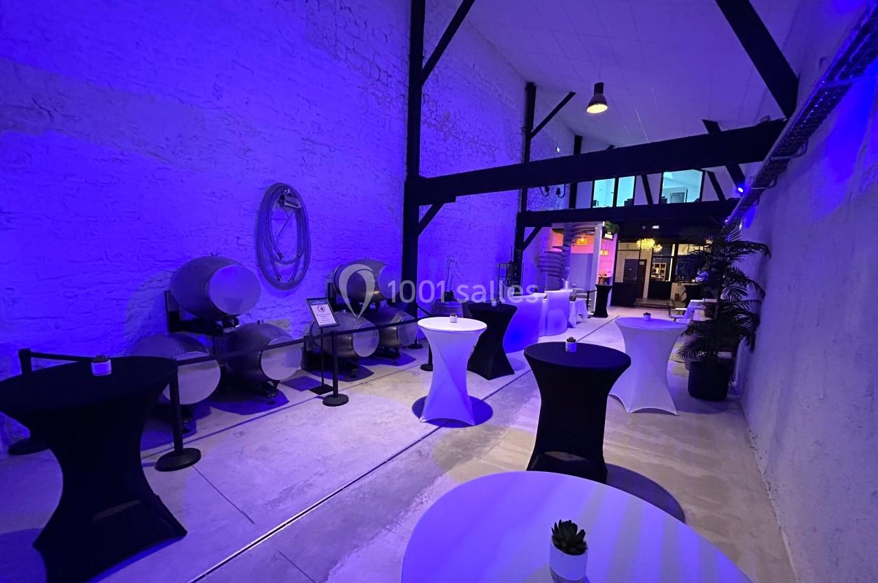 Salle avec murs en pierre, tonneaux en arrière-plan et tables hautes éclairées par une lumière bleue.