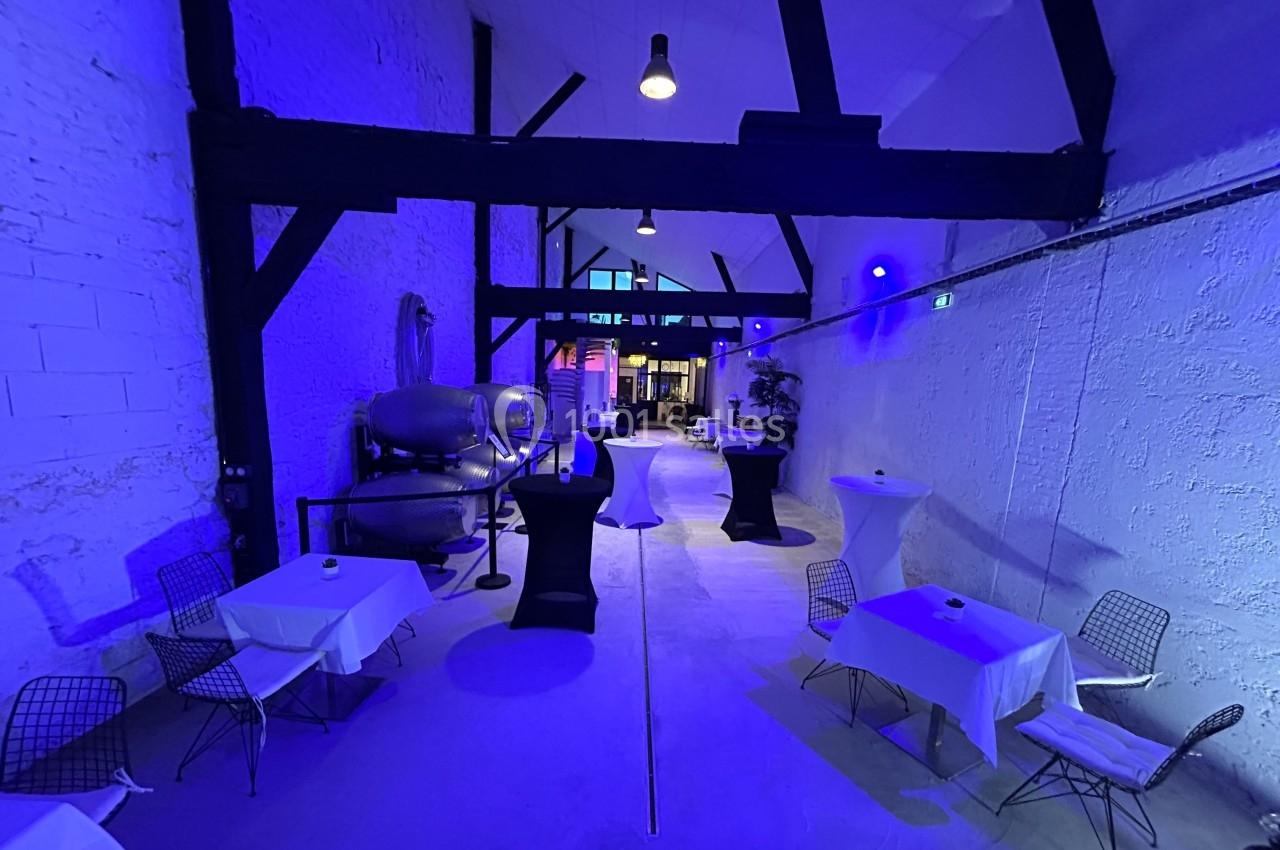 Salle industrielle avec murs blancs, éclairage bleu, tables dressées et chaises modernes, ambiance épurée et contemporaine.