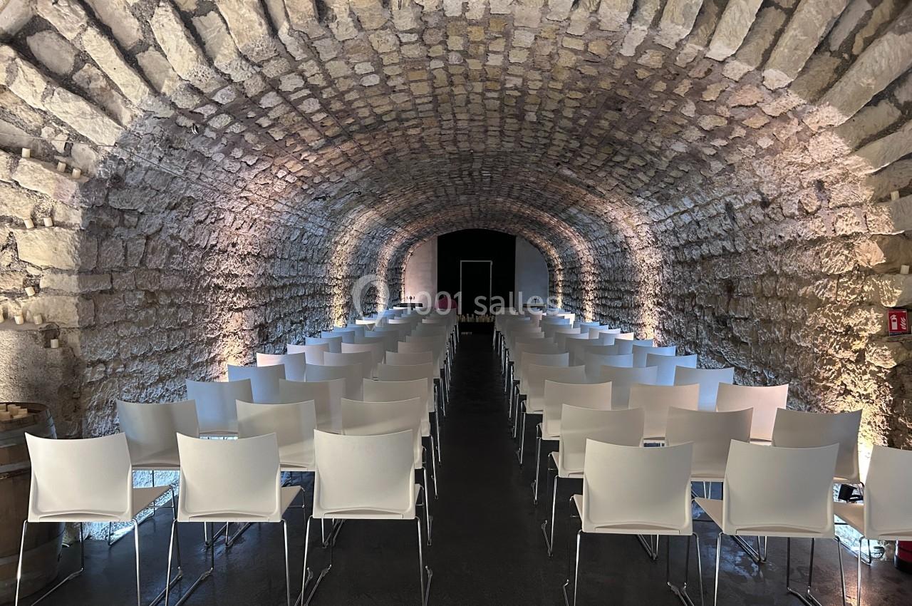 Salle voûtée en pierre aménagée avec des rangées de chaises blanches alignées face à un espace de présentation.
