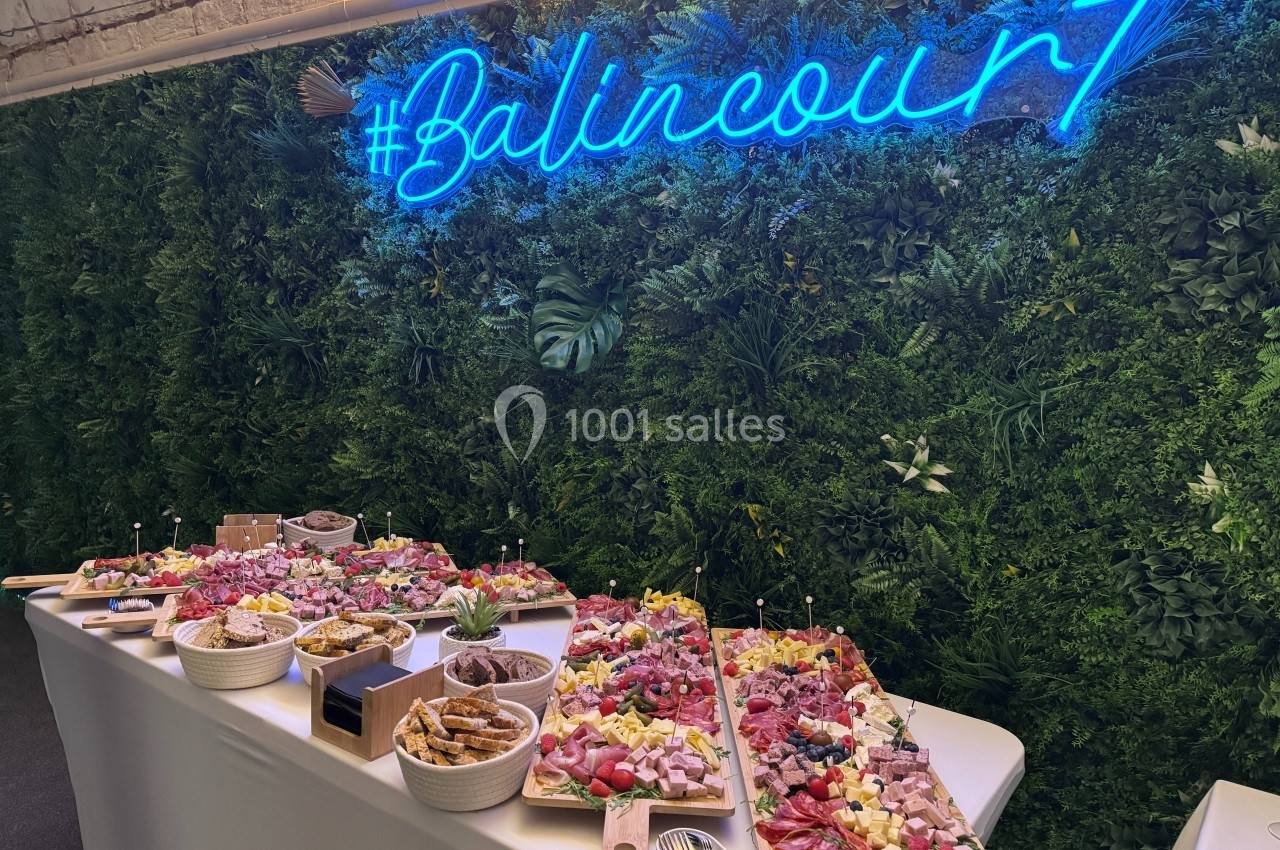 Buffet garni de planches de charcuterie, fromages et fruits, disposé devant un mur végétal avec un néon bleu.