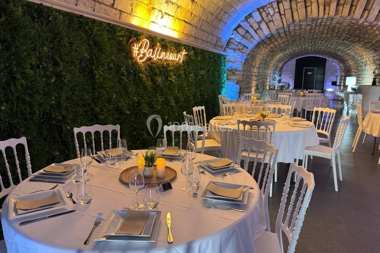 Salle voûtée décorée avec des tables rondes dressées, nappes blanches, mur végétal et enseigne lumineuse.