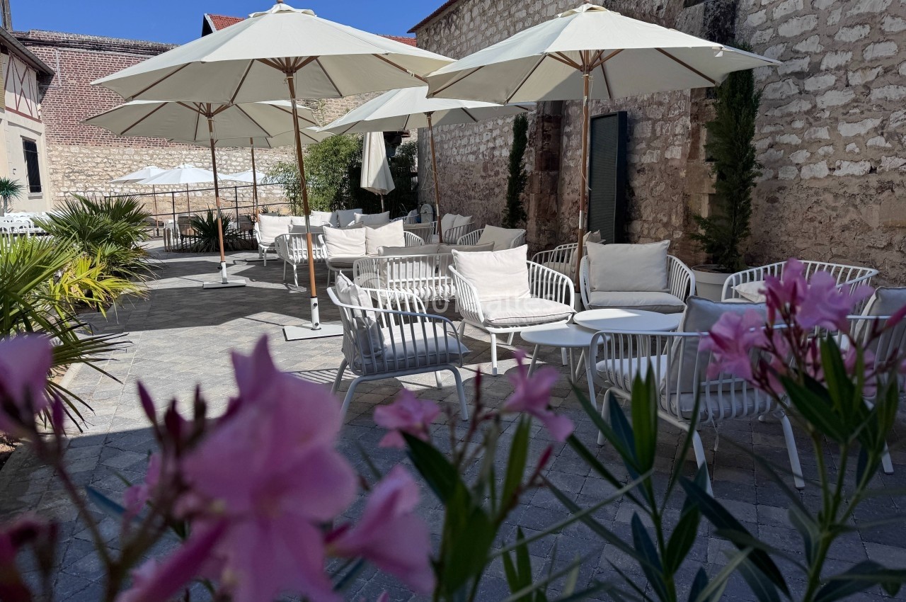 Terrasse ensoleillée avec tables, chaises blanches et parasols, entourée de murs en pierre et de fleurs roses au premier…