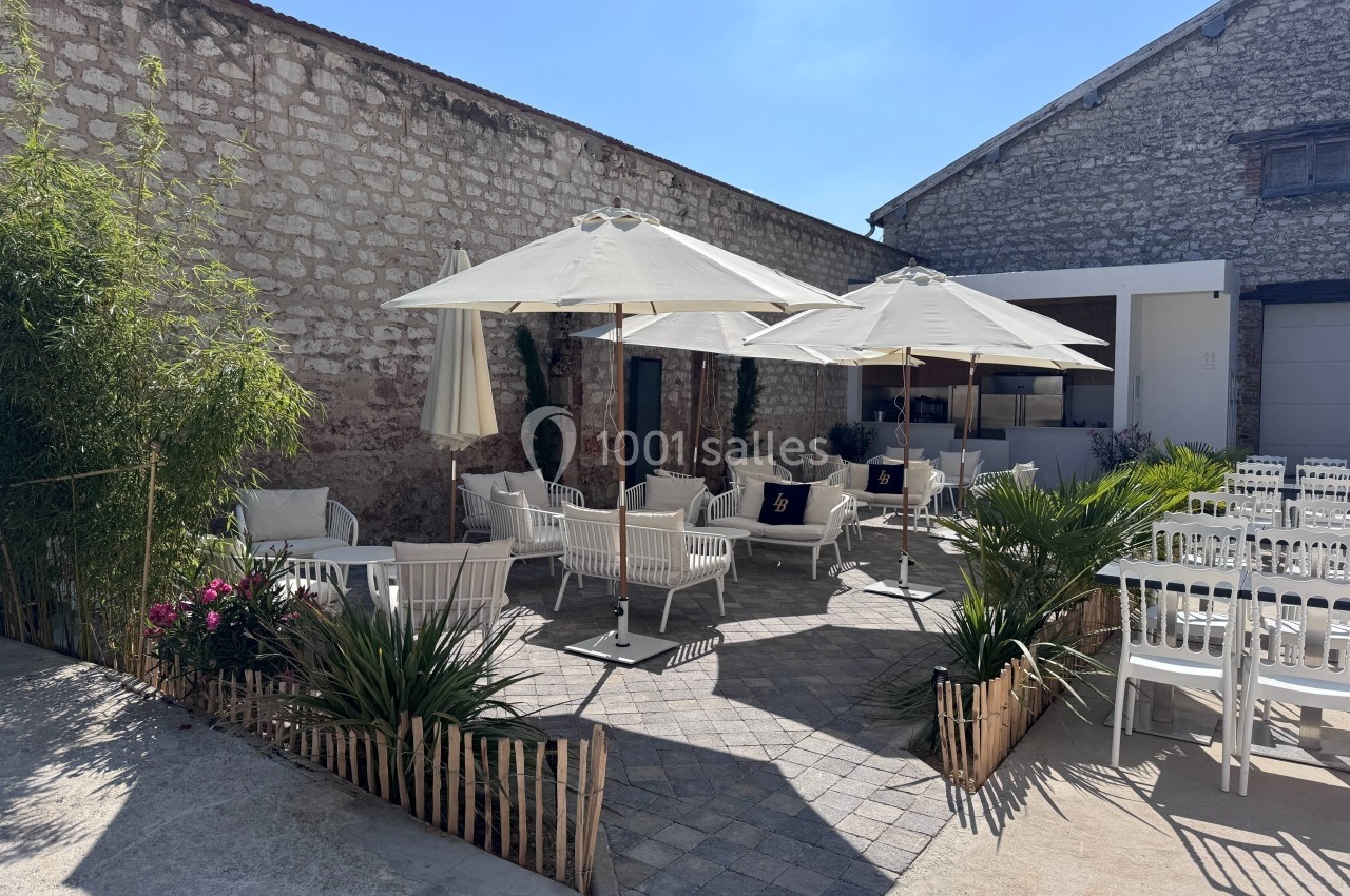 Terrasse extérieure avec mobilier blanc, parasols et végétation, entourée de murs en pierre sous un ciel ensoleillé.
