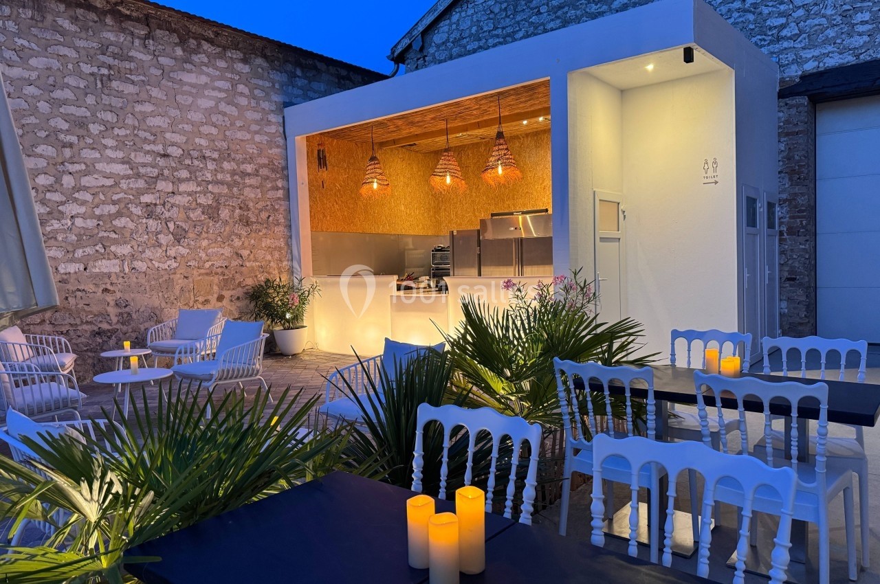 Terrasse extérieure en soirée avec tables, chaises blanches, bougies allumées et un comptoir éclairé en arrière-plan.