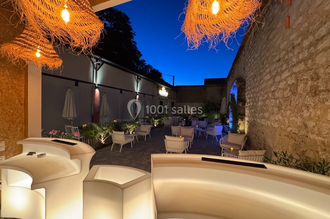 Terrasse extérieure d'un restaurant en soirée, éclairée par des lampes suspendues et entourée de murs en pierre.