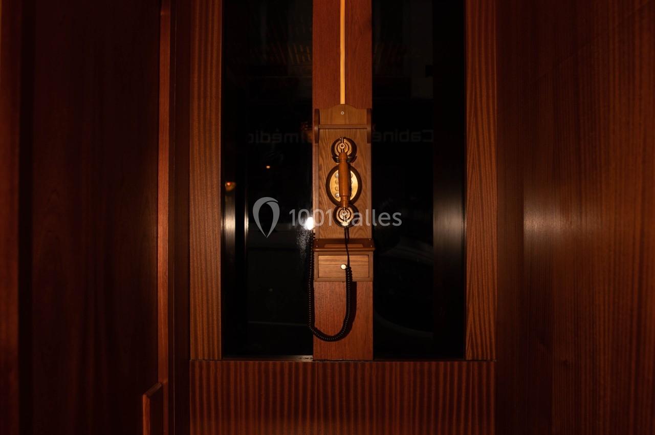 Cabine d'ascenseur en bois sombre avec un téléphone mural rétro et une lumière décorative au plafond.