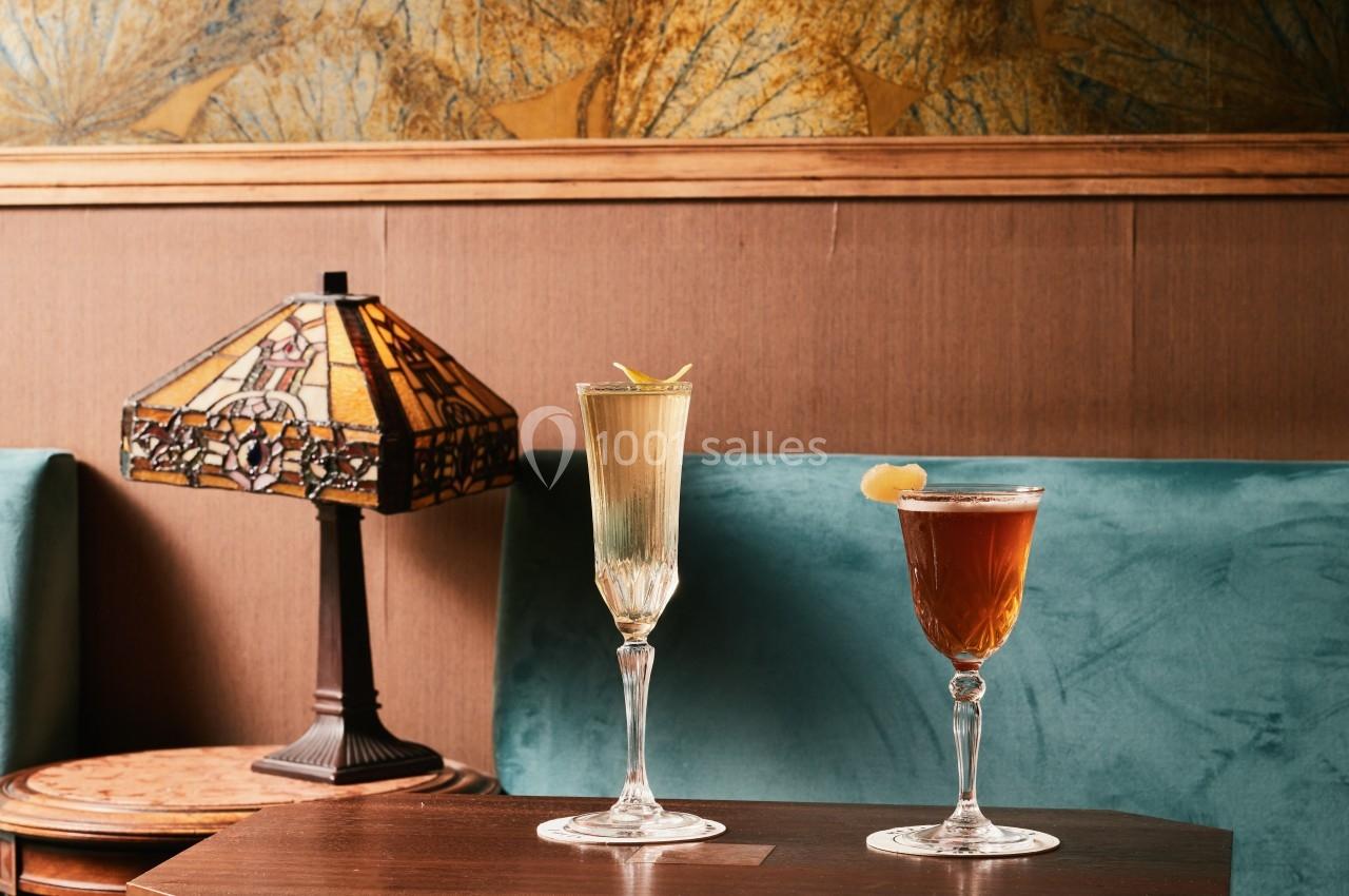 Deux verres de cocktails élégants posés sur une table en bois, avec une lampe vintage et un canapé en arrière-plan.