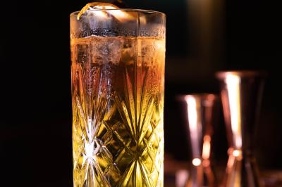 Une main tenant un verre de vin blanc dans un environnement sombre, avec des personnes et une lampe en arrière-plan.