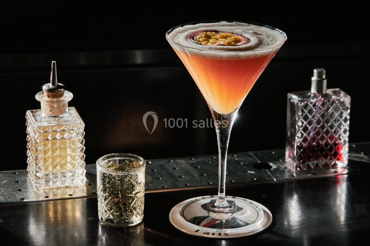 Cocktail dans un verre à martini garni d'un demi-fruit de la passion, entouré de flacons sur un bar sombre.