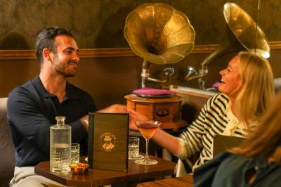 Une main tenant un verre de vin blanc dans un environnement sombre, avec des personnes et une lampe en arrière-plan.