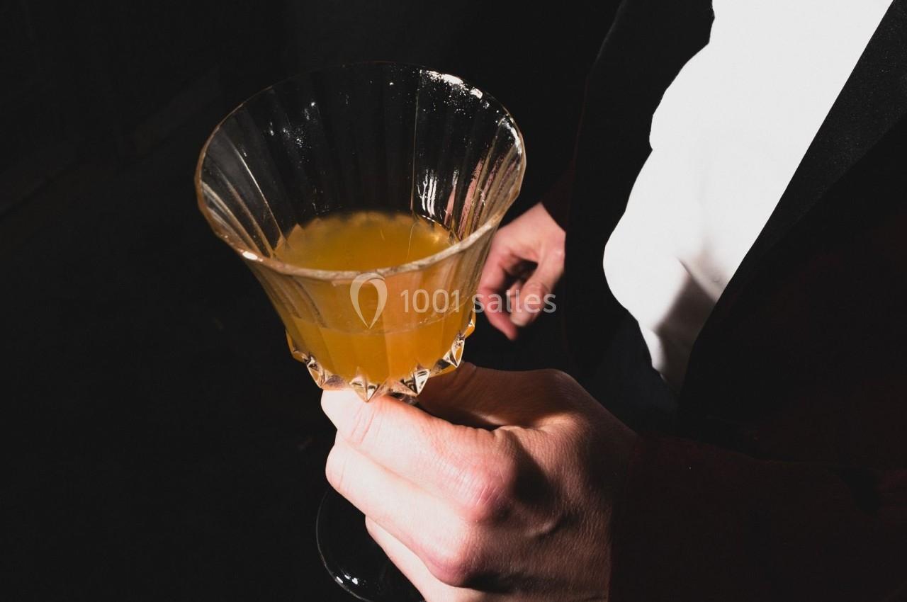 Main tenant un verre à cocktail rempli d'une boisson jaune, éclairé par une lumière contrastée dans un environnement sombre.