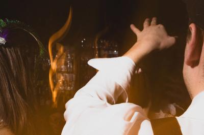 Une main tenant un verre de vin blanc dans un environnement sombre, avec des personnes et une lampe en arrière-plan.