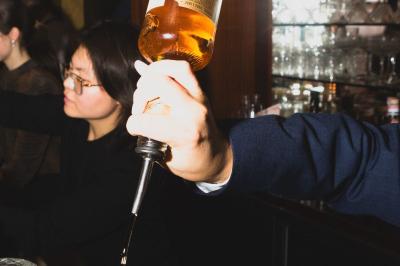 Une main tenant un verre de vin blanc dans un environnement sombre, avec des personnes et une lampe en arrière-plan.