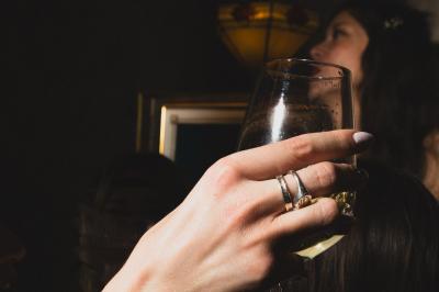 Une main tenant un verre de vin blanc dans un environnement sombre, avec des personnes et une lampe en arrière-plan.