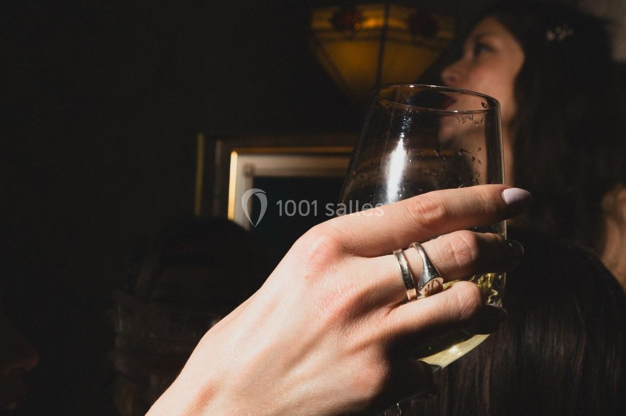 Une main tenant un verre de vin blanc dans un environnement sombre, avec des personnes et une lampe en arrière-plan.