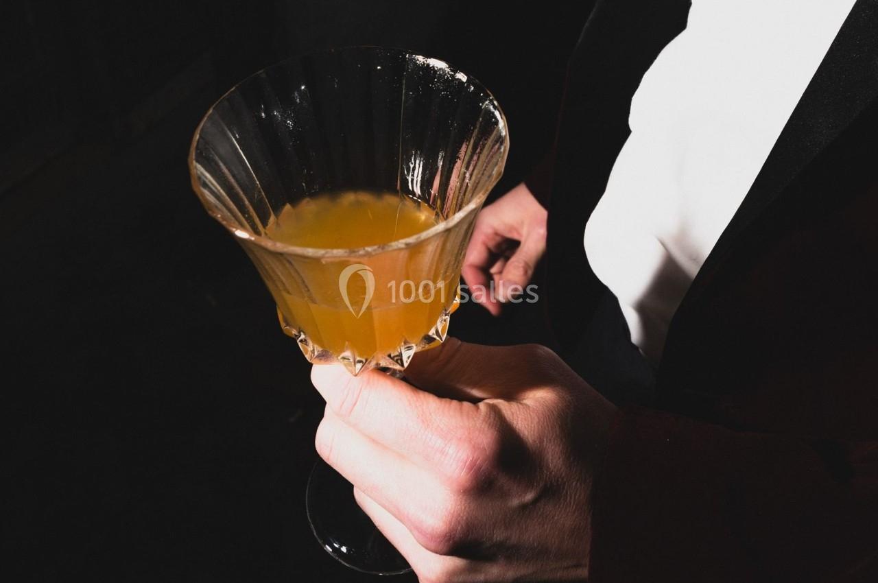 Une main tenant un verre à cocktail rempli d'une boisson orange, sur un fond sombre.